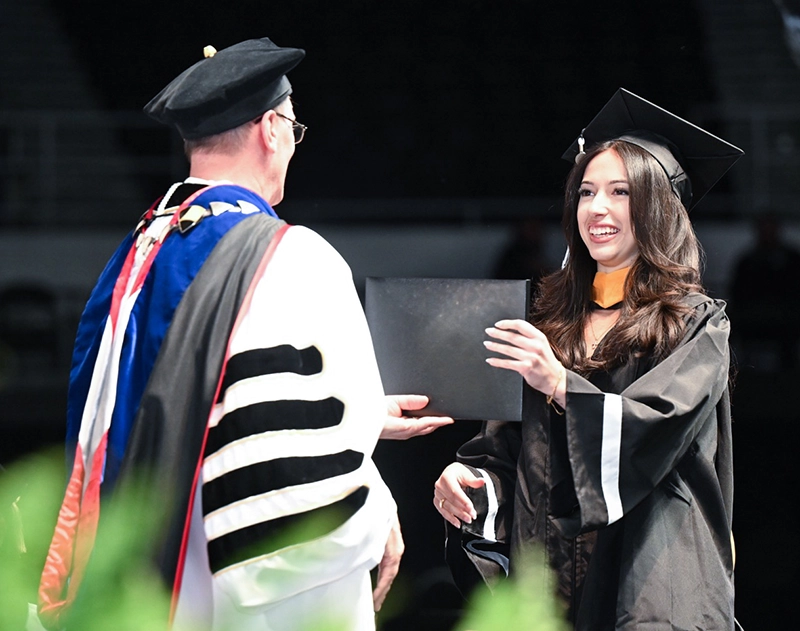 A young woman in black cap and gown accepts a diploma from the college president with a large smile