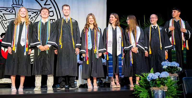 Eight students in caps and gowns stand on a stage