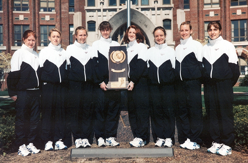 Eight women wearing track suits pose in front of a brick building holding a plaque from the NCAA cross country championship.