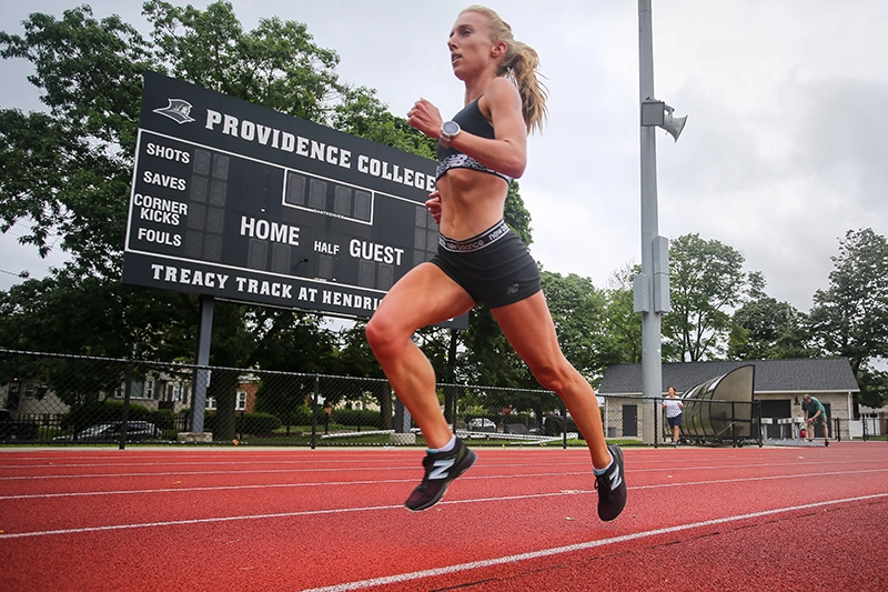 A woman dressed in black running gear runs on a red turf track alongside a scoreboad.