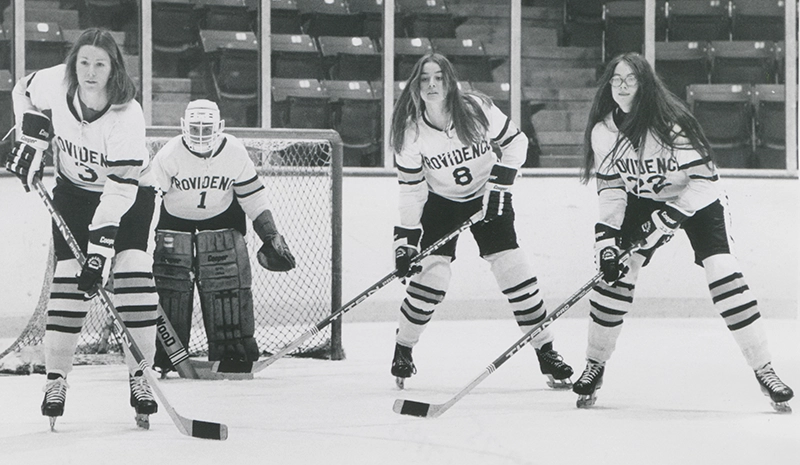 Women's players stand in front of the net with sticks, awaiting the puck.