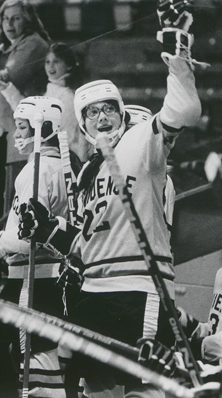 A women's hockey player in a helmet waves a mitt toward the crowd in greeting.