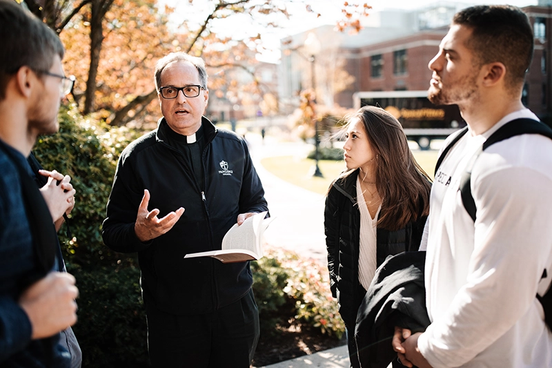 A priest talks to three students on a path with brick college buildings in the background.