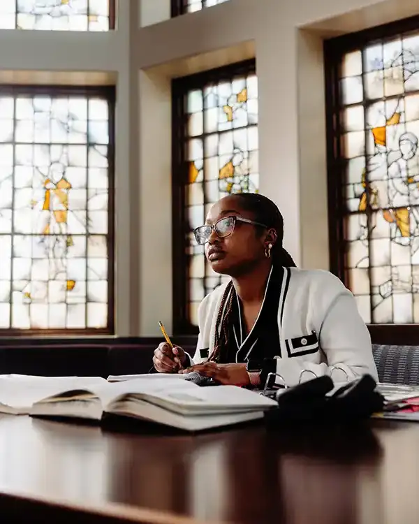 Angie Pierre '25 sits at a table in the Fiondella Great Room of the Ruane Center for the Humanities at Providence College. She is writing with a pencil and there are books open on the desk in front of her. There are stained glass windows behind her.