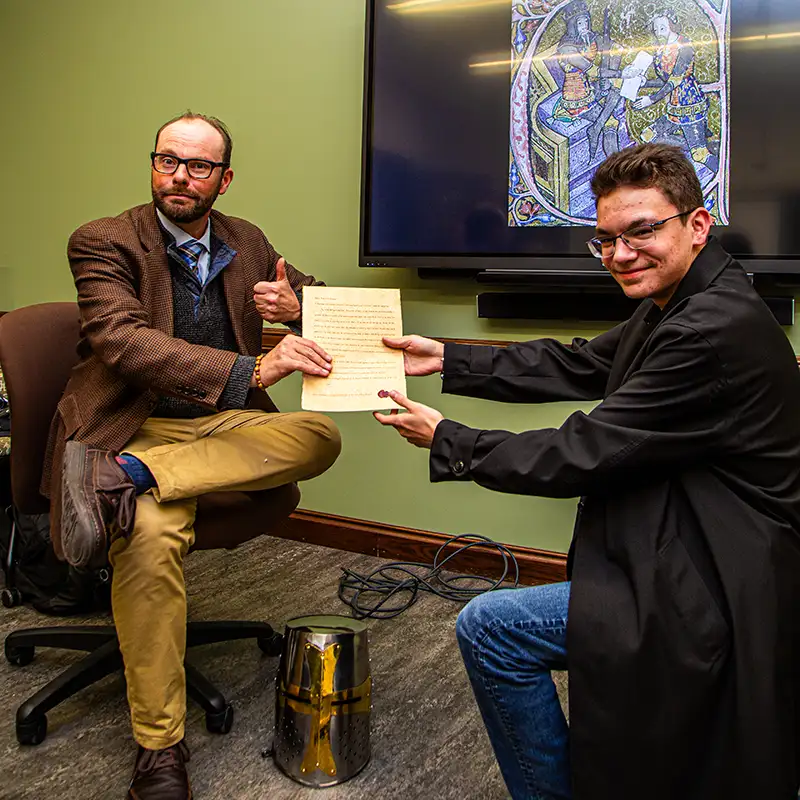 A student hands Berard his assignment mimicking an Arthurian scene displayed on the screen behind