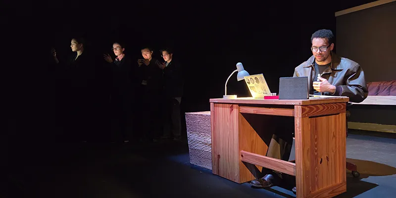 Rodney Lopez as Thomas Stockmann at his desk. To the side of the stage are other cast members illuminated by the screens of their phones.