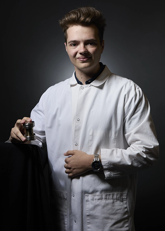 A young man in a lab coat holds a brass cylinder.