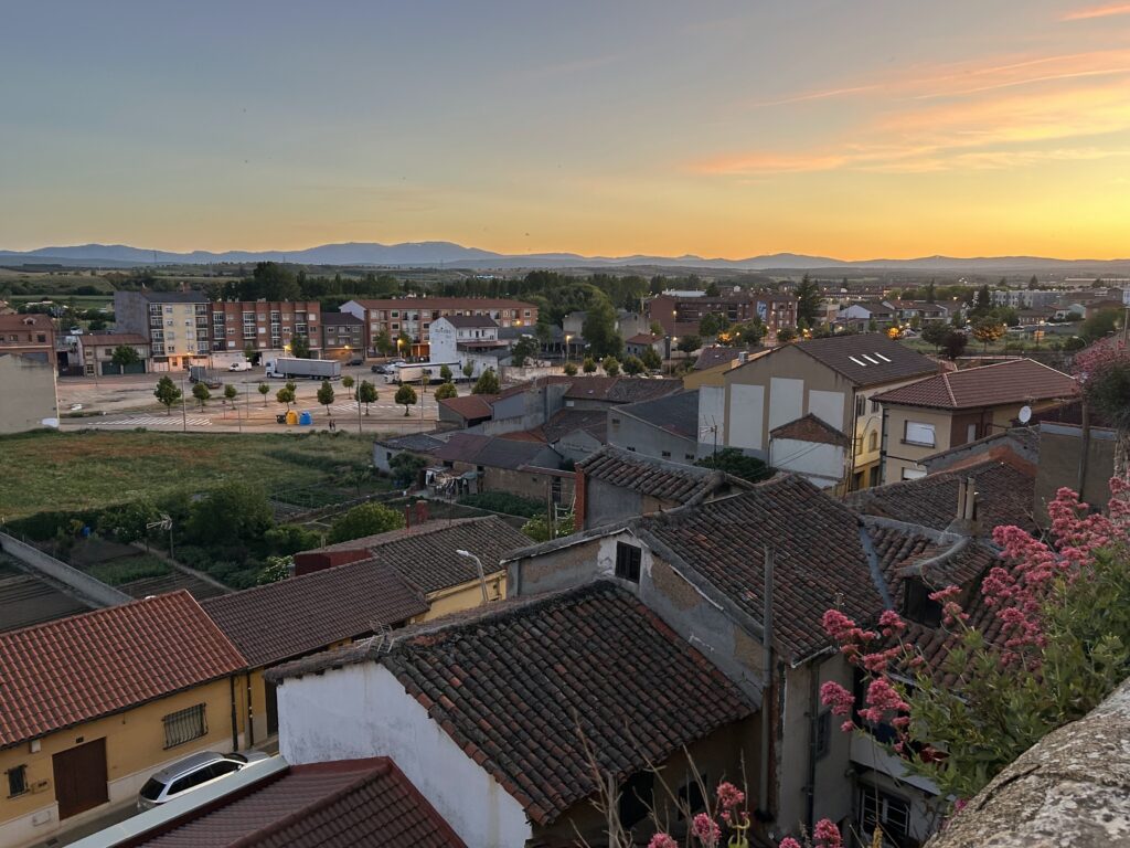 A scenic view of a small town at sunset, with rooftops in the foreground and a backdrop of distant mountains. The sky is tinged with warm orange and pink hues.