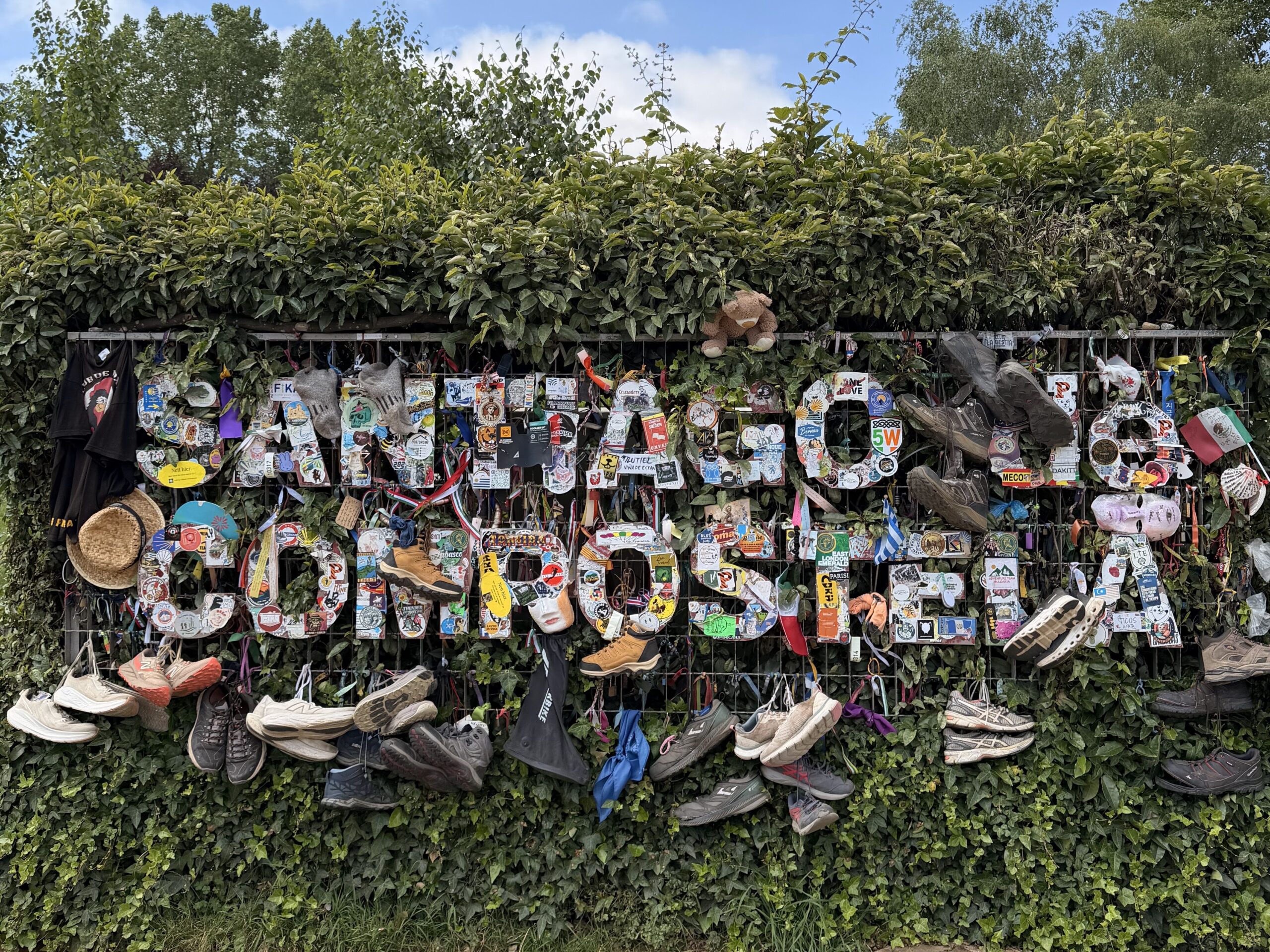 The image shows a green hedge with a metal frame in front, adorned with numerous shoes, hats, and small items. The frame spells out "Santiago de Compostela." The ground is dirt, and the sky is partly cloudy.