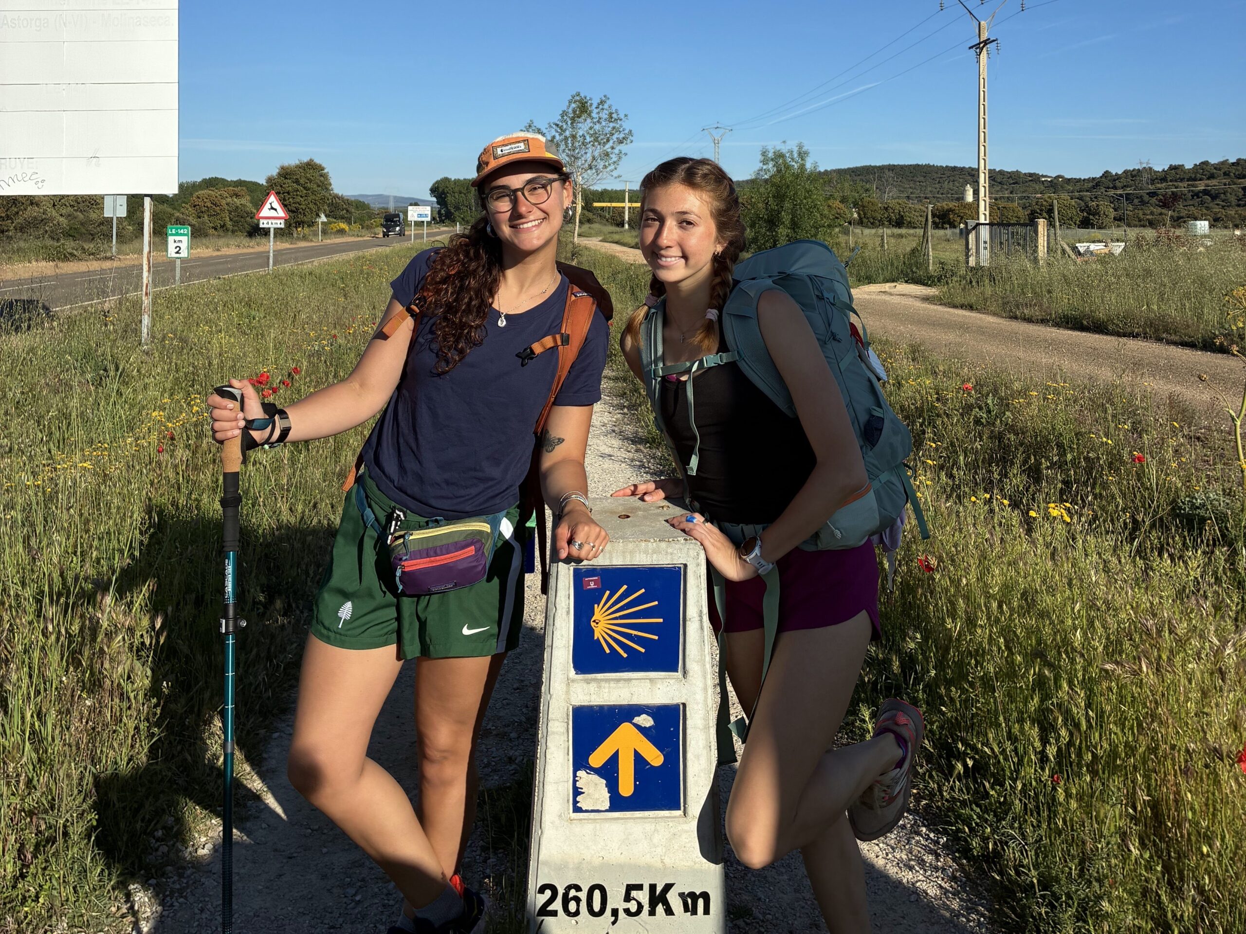 Two people are smiling and standing next to a Camino de Santiago marker that reads "260,5 KM" with a yellow arrow and shell symbol. They are wearing backpacks and holding walking poles on a sunny day, with a road and countryside in the background.