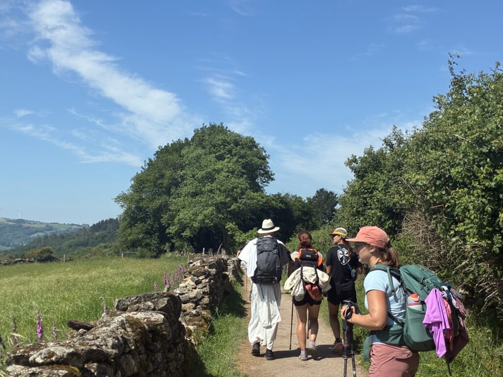 A group of people are walking on a country trail surrounded by stone walls and greenery. The sky is clear with wispy clouds. The path extends toward distant trees.