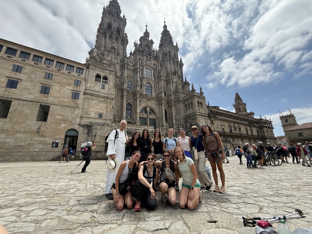 A group of people posing in front of the Santiago de Compostela Cathedral, under a partly cloudy sky. Some are kneeling in front, while others stand behind. The cathedral's intricate architecture is visible in the background, with several other people walking around the plaza.
