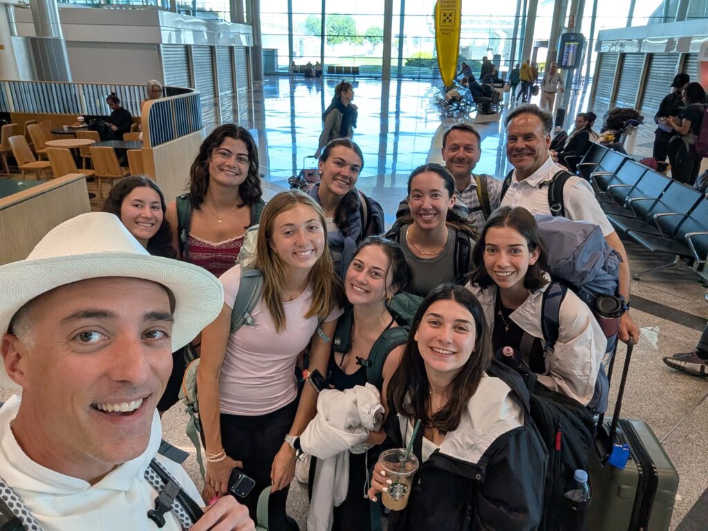 A group of people smiling and posing for a selfie in an airport terminal. They are surrounded by chairs and other travelers in the background. Everyone appears prepared for travel with backpacks and luggage.