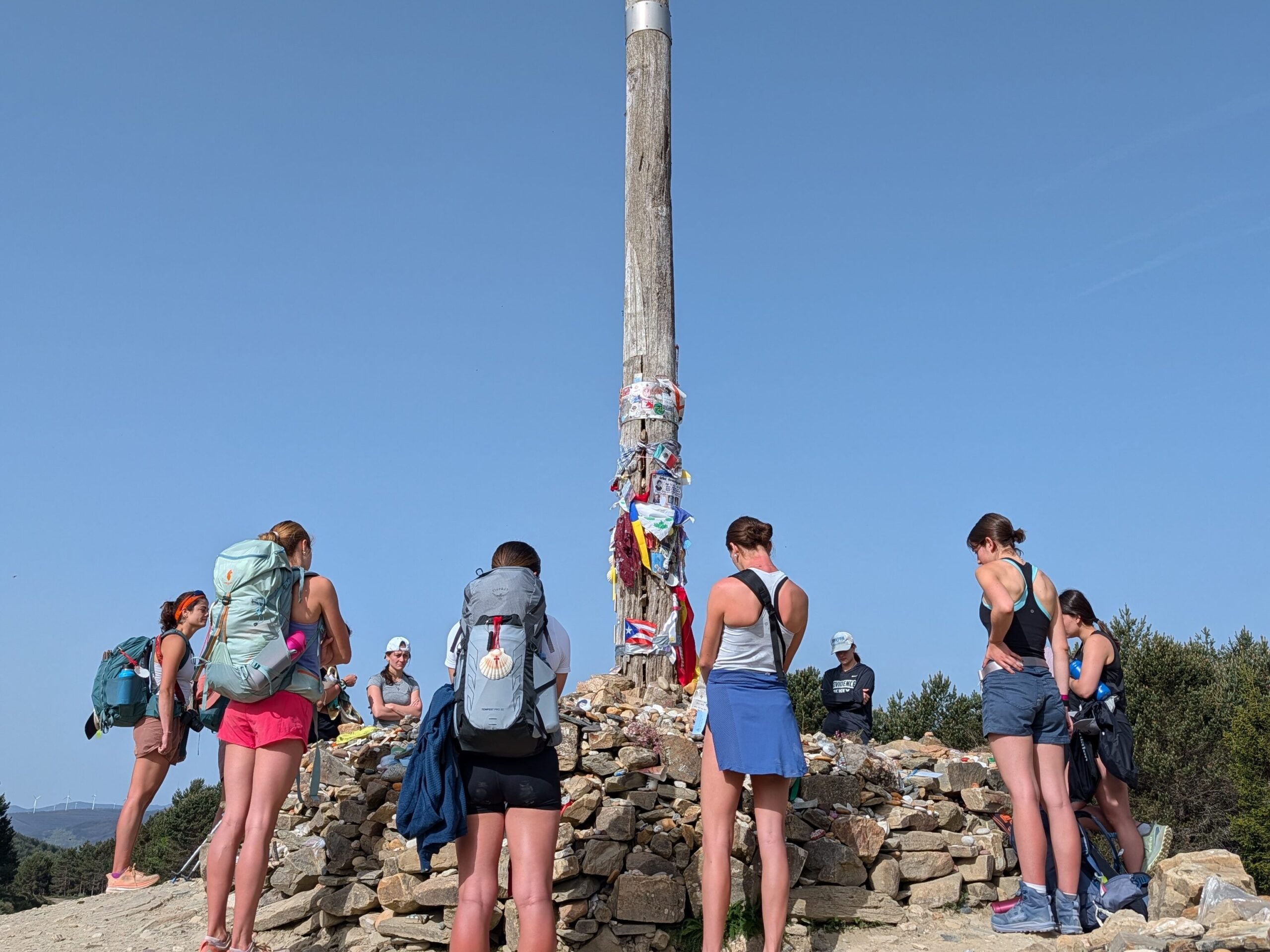 A group of people stand around the Iron Cross, a tall wooden pole adorned with ribbons and mementos on top of a pile of stones, along the Camino de Santiago.