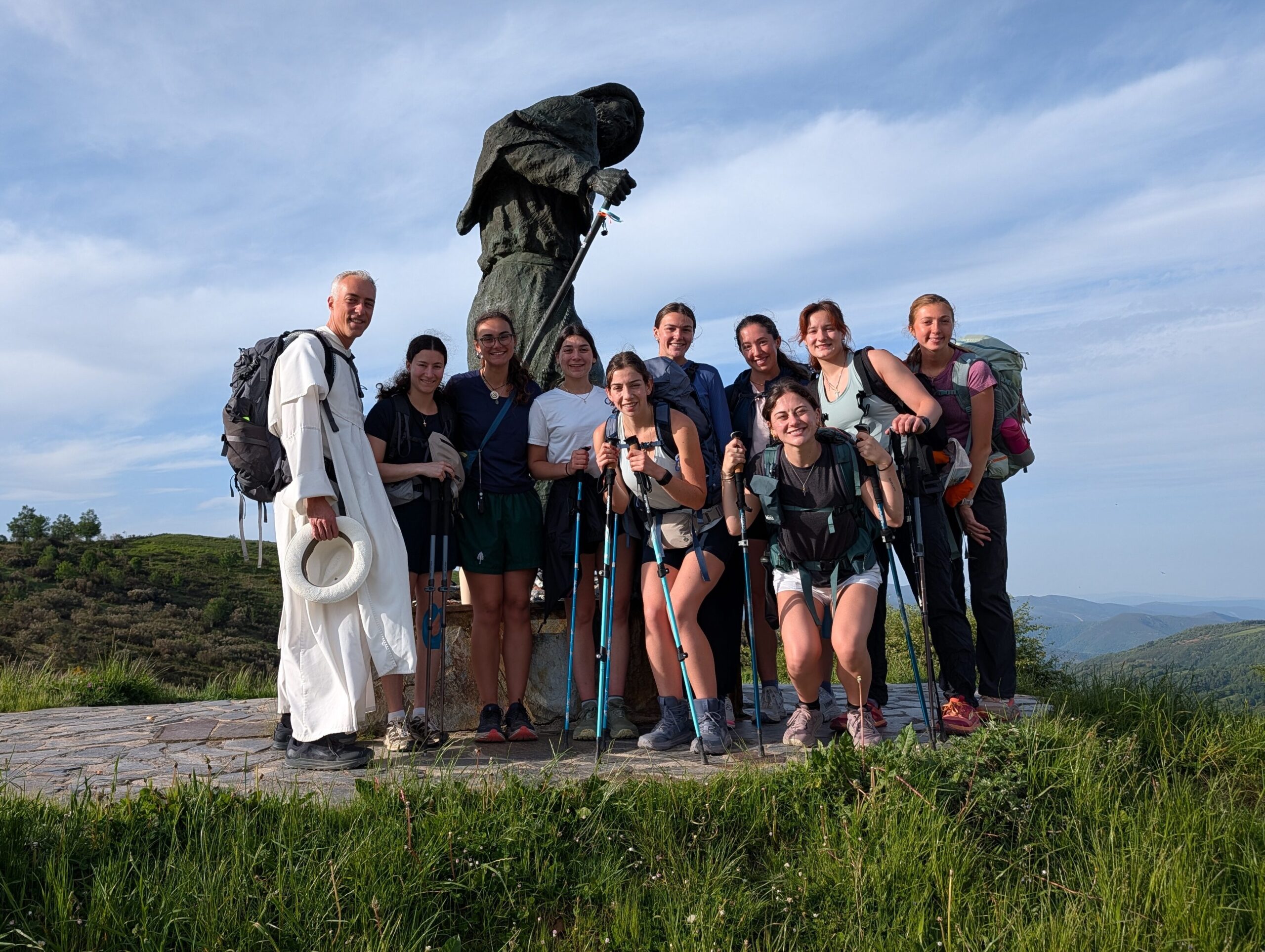 A group of people with backpacks and hiking poles pose in front of a large statue on a grassy hill. The sky is clear with a few clouds in the background.