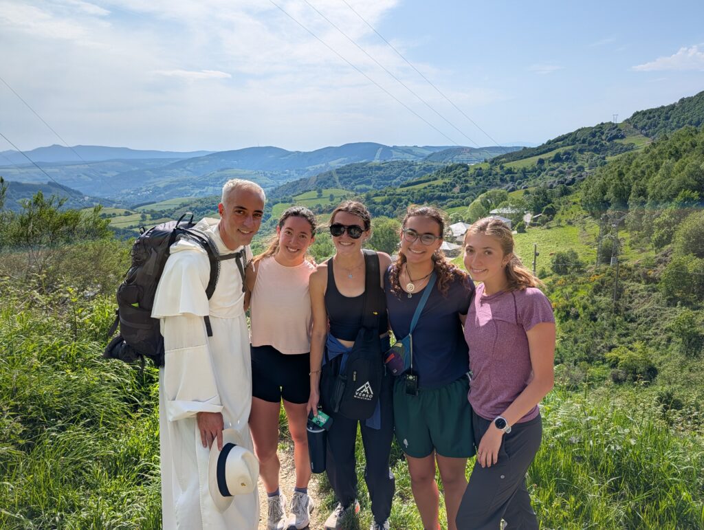 A group of five people smiling, standing on a scenic trail with rolling green hills and forests in the background. One person is wearing a white robe and carrying a backpack. It's a sunny day with a clear blue sky.